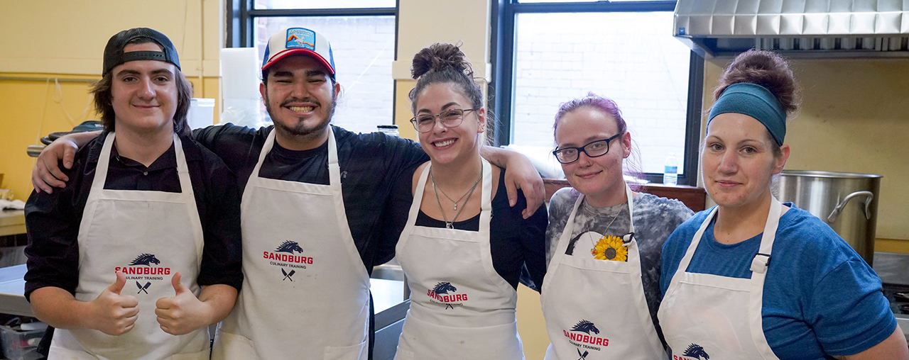 Five people standing in a commercial kitchen with aprons on smiling.