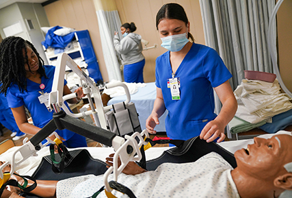 CNA student in the nursing lab looking at a hospital bed with a sim man in it.