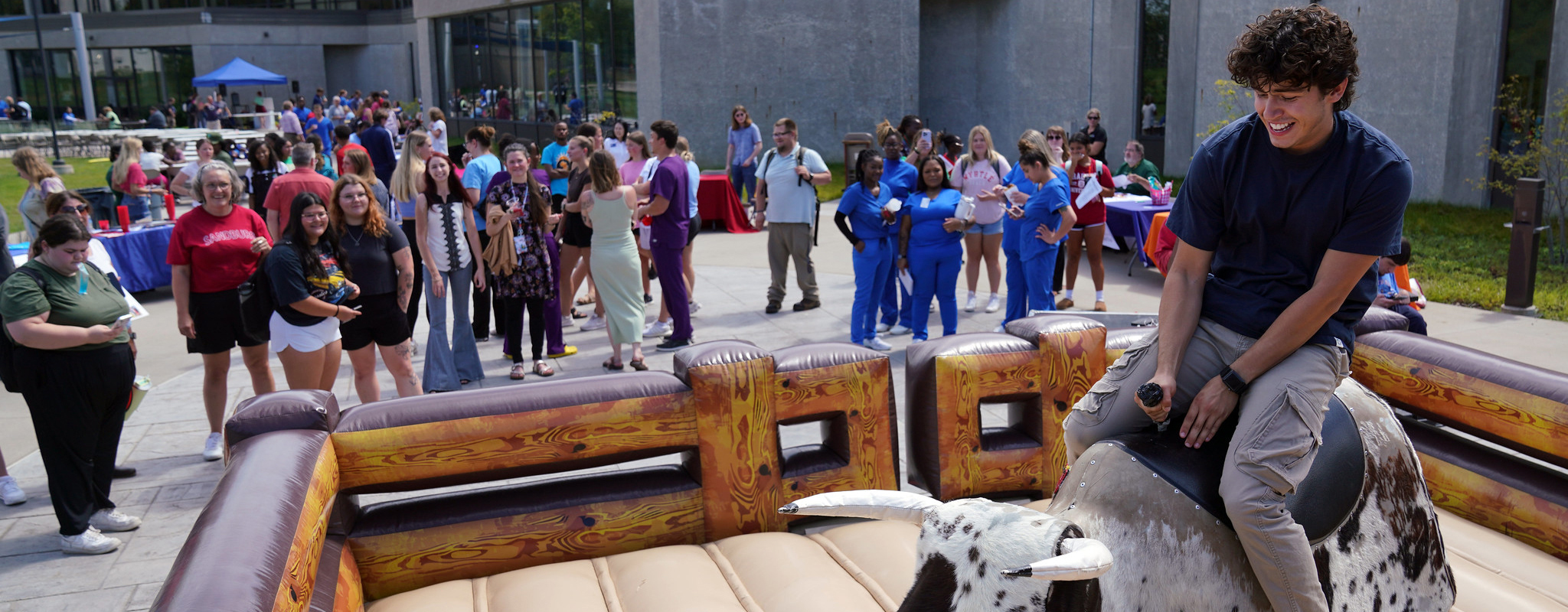 Male student riding a mechanical bull as other students watch.